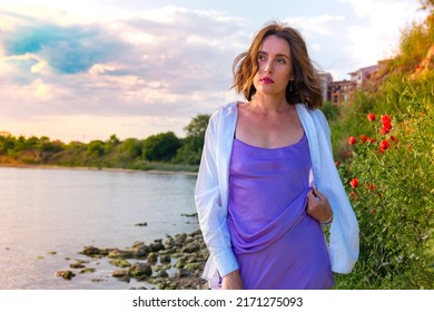 A Beautiful Young Girl In A White Shirt And Purple Dress Poses Against The Backdrop Of A Pond And Blooming Poppies. Photo Session Of A Beautiful Young Woman At Sunset.