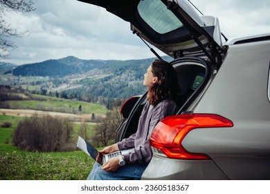 A beautiful young girl is working on a laptop in the trunk of a car and admiring the views of the mountains while traveling through the highlands. Remote work, downshifting, work and travel. - Powered by Shutterstock