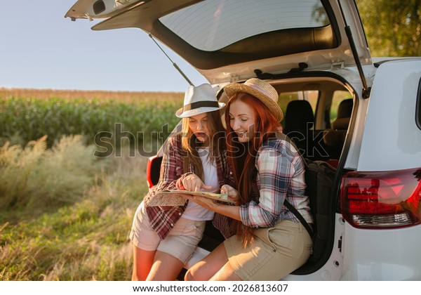  Beautiful\
young girls seating in the  car trunk with backpack and luggage\
wearing white hat checking road map on paper map, excited ready for\
road trip,  corn field on\
background