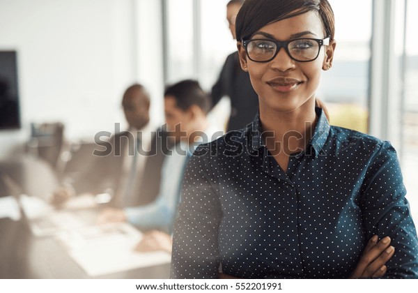 Beautiful young grinning\
professional Black woman in office with eyeglasses, folded arms and\
confident expression as other workers hold a meeting in\
background