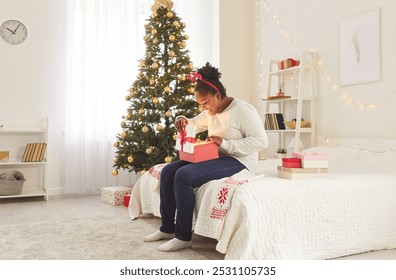 Beautiful young happy african american woman opening red present gift box sitting on cozy bed in bedroom with fir tree at home. Joyful excited girl sitting on Christmas eve or celebrating New Year. - Powered by Shutterstock