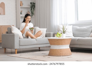 Beautiful young happy Asian woman with bowl of fresh vegetable salad sitting on sofa in living room - Powered by Shutterstock