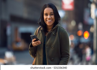 Beautiful young happy latin woman texting on mobile phone on city street. Student girl walking and texting on cell phone outdoors on city street at winter time.