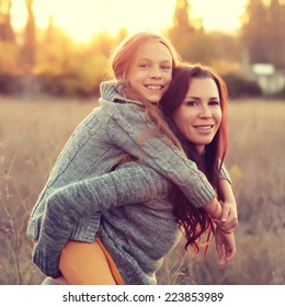 Beautiful Young Happy Mother With Little Girl On The Back. Closeup Of A Happy Family On A Walk In A Field In The Sunshine. Photo Toned Style Instagram Filters.