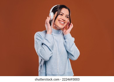 Beautiful Young Happy Smiling Brunette Woman Girl In Blue Sweater, Listens Music With Headphones, Isolated On Brown Wall Background Studio Portait. People Lifestyle Concept. Mock Up Copy Space.