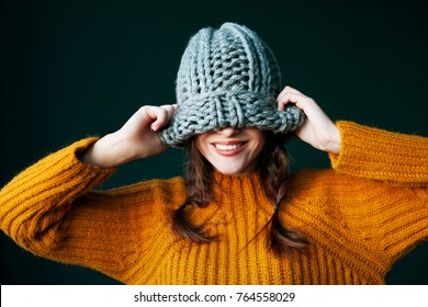 Beautiful young happy smiling girl hiding her eyes under trendy grey big loop knitted beanie hat. Model wearing winter sweater. Close up