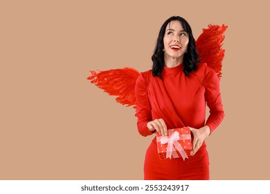 Beautiful young happy woman dressed as cupid with gift box on beige background. Valentine's Day celebration
