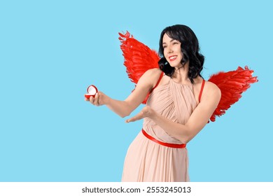 Beautiful young happy woman dressed as cupid with engagement ring on blue background. Valentine's Day celebration