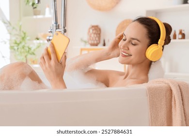 Beautiful young happy woman in headphones holding mobile phone and taking bath with soap foam at home - Powered by Shutterstock