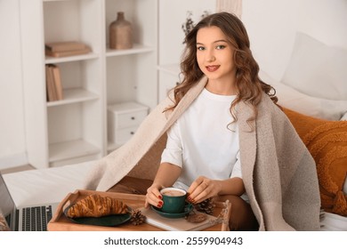 Beautiful young happy woman with plaid, cup of coffee, laptop and croissant on wooden tray sitting at home - Powered by Shutterstock