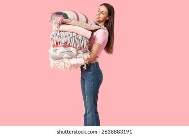Beautiful young happy woman with stack of different blankets and pillow on pink background