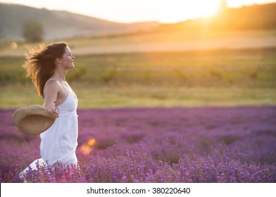 Beautiful young  healthy woman with a white dress running joyfully through a lavender field holding a straw hat under the rays of the setting sun.