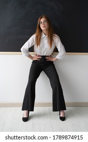 Beautiful Young High School Teacher In The Office. She Is Standing In Front Of A Clean Black Board. She Is Wearing A White Women's Shirt And Black Trousers, As Well As Black Pointed Toe Shoes. Her