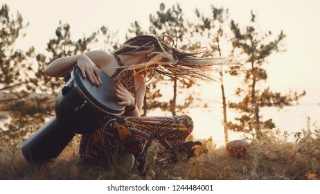 Beautiful young hippie woman with dreadlocks playing on djembe. Funky woman drumming in nature on an ethnic drum at sunset or sunrise - Powered by Shutterstock