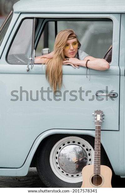 A\
beautiful young hippie woman in yellow sunglasses looks out the\
window of a blue vintage minivan. Make love, not war. Girl child\
flowers driving in a retro van. Auto travel\
concept.