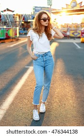 Beautiful young hipster girl in sunglasses in a white T-shirt, vintage jeans and white sneakers walking in the amusement park at sunset.