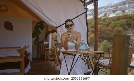 A beautiful young hispanic brunette woman wearing sunglasses and a white dress sits at a table in an outdoor camping tent setup in africa, using her smartphone. - Powered by Shutterstock