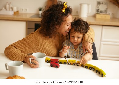 Beautiful Young Hispanic Mom Embracing Adorable Baby Boy, Kissing Him On Forehead, Having Morning Coffee, Sitting At Kitchen Table, Playing With Toys Together In The Morning. Family And Generations