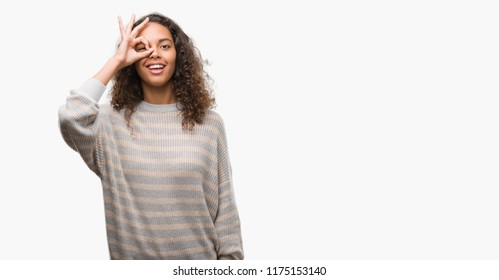 Beautiful young hispanic woman wearing stripes sweater doing ok gesture with hand smiling, eye looking through fingers with happy face.