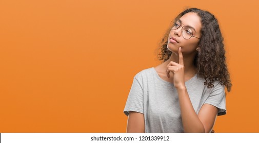 Beautiful young hispanic woman wearing glasses with hand on chin thinking about question, pensive expression. Smiling with thoughtful face. Doubt concept.