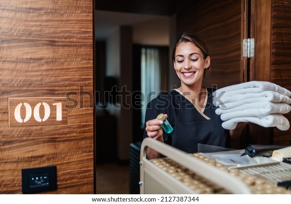 Beautiful young hotel chambermaid\
in uniform bringing clean towels and other supplies to hotel\
room.