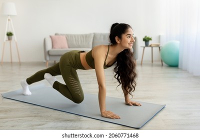 Beautiful young Indian woman doing strength exercises, training her abs muscles on sports mat at home, full length. Pretty Asian lady training in living room. Domestic fitness during coronavirus
