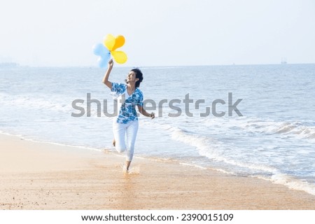 Beautiful young indian woman running with multicolor balloon in hands at the beach. Young girl having fun, holidays and vacation.