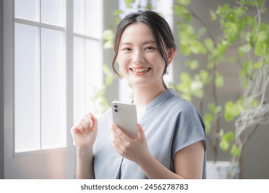 A beautiful young Japanese woman with a smartphone stands by the window of a white office. She is smiling and looking at the camera. She taps the screen of her smartphone. - Powered by Shutterstock