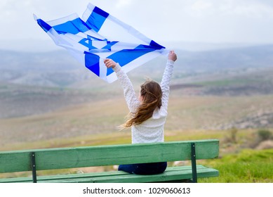Beautiful young jewish girl sitting holding  Israel flag in the wind and  enjoying great view landscape on the sky, field and mountains.Patriotic holiday. Independence day Israel - Yom Ha'atzmaut .