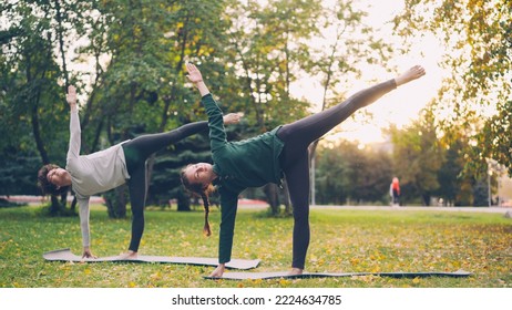 Beautiful young ladies are standing in Half Moon pose Ardha Chandrasana during pair yoga class in park in autumn. Healthy lifestyle, teaching and learning and sports concept. - Powered by Shutterstock