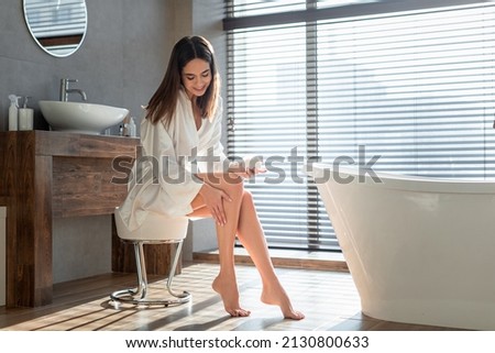 Beautiful young lady applying moisturising lotion on legs while sitting in bathroom interior, smiling attractive female rubbing nourishing cream, doing skincare treatments at home, copy space