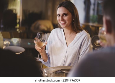 Beautiful young lady carefree enjoying a glass of white wine in an elegant restaurant on a weekend night sitting at a table with her friends - people, alcohol, lifestyle concept
