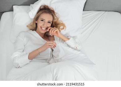beautiful young lady with light hair relaxing in wide and comfortable bed with snow-white pillows and blanket. Smile joyful , looking happily , dreaming about something funny and cute. 