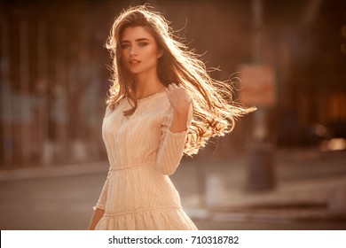 Beautiful young lady with long healthy hair and cute dress walking on the street. Hot summer evening. Sunset. Lifestyle concept.