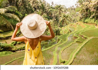 Beautiful young lady in shine through dress touch straw hat. Girl walk at typical Asian hillside with rice farming, mountain shape green cascade rice field terraces paddies. Ubud, Bali, Indonesia.