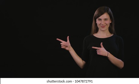A beautiful young lady smiling and standing and pointing with her fingers against a black background. Medium Shot