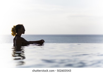 Beautiful Young Lady Swimming In An Infinity Pool Wearing A Flower Hat And Watching The Sun Set