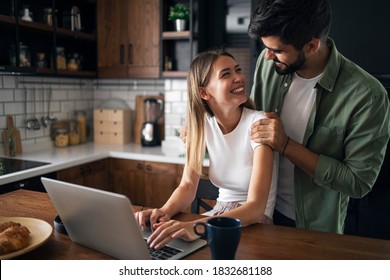 Beautiful Young Lady Working From Home On Laptop. Man Is Smiling At Her. Happy Couple Enjoying Work From Home