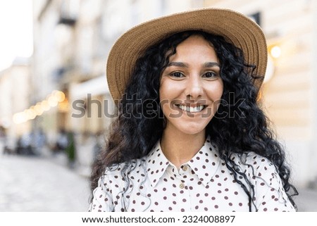 Beautiful young Latin American woman portrait, woman walking in evening city in hat with curly hair in warm weather, smiling and looking at camera close up.