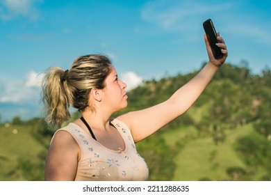 Beautiful Young Latin Woman On Top Of A Colombian Mountain Raising Her Hand To Look For A Signal For Her Mobile Phone. Girl Looking For Satellite Reception On Top Of A Hill. Satellite Internet Concept