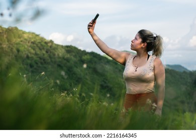 Beautiful Young Latina Woman, On Top Of A Mountain Looking For Network Coverage For Her Cell Phone. Girl Looking For Satellite Signal, Hiking On Top Of A Hill. Technology Concept