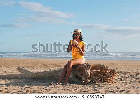 A beautiful young latina woman in yellow swimsuit drinking coconut water sitting on a log on the beach