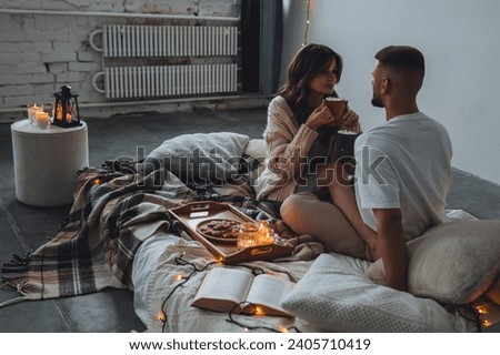 Beautiful young loving couple having breakfast, drinking hot chocolate or coffee with marshmallow in bed at home. Happy spouses enjoying lazy romantic winter morning in bedroom. Candles, garlands