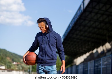 Beautiful young male model wearing a blue hoodie and headphones holding the ball on the court smiling and looking down - Powered by Shutterstock