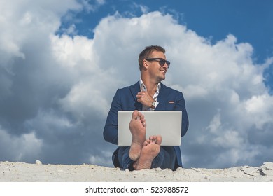 Beautiful Young Man Working On Laptop While Sitting Barefoot On Sand In Desert Enjoying Nature And The Sun. Peaceful Place To Work And Leisure.