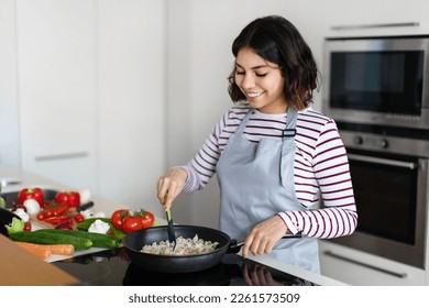Hermosa joven mujer de medio oriente sosteniendo espátula de cocina, mezclando arroz con verduras en sartén en estufa eléctrica, preparando comida en la cocina, espacio de copia. Dieta saludable, nutrición