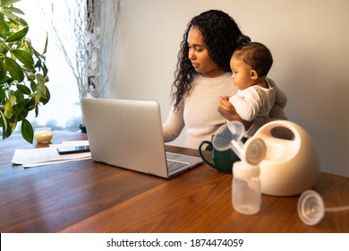 A beautiful young mixed Race African American mother holds her daughter while taking notes at her dining table serving as a temporary remote work from home station with breast pump in foreground.