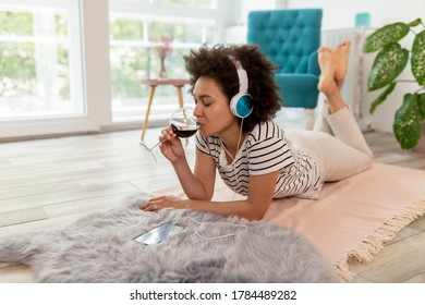 Beautiful Young Mixed Race Woman Lying On The Living Room Floor, Listening To The Music, Drinking Wine And Enjoying Leisure Time At Home