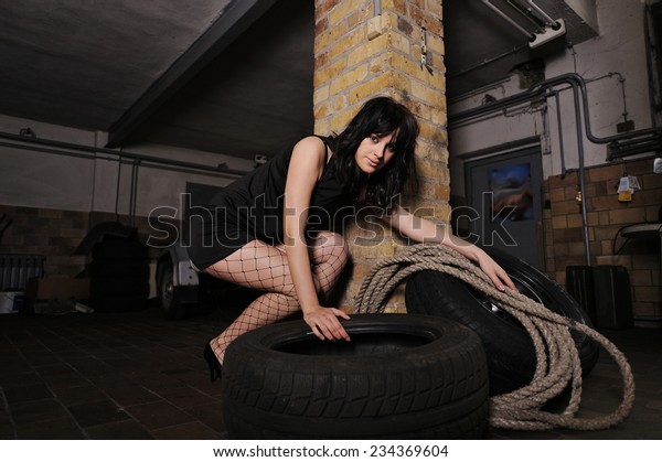 A beautiful\
young model in a car service. She is picking up a dirty wheel while\
holding a rope in the other\
hand.