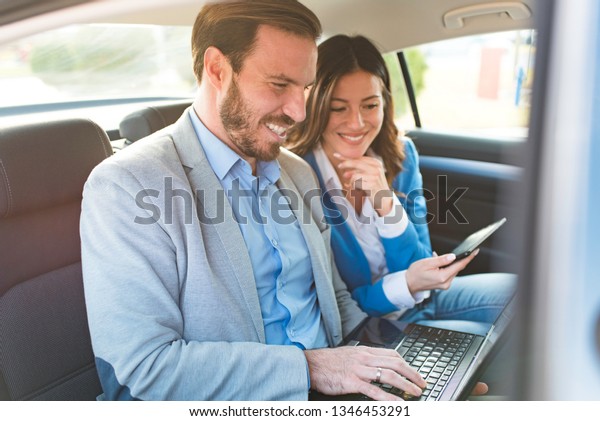 Beautiful\
young modern business people driving in the backseat of a luxury\
car using laptop and phone, working together and smiling in the car\
while driving to work wearing elegant\
suits.